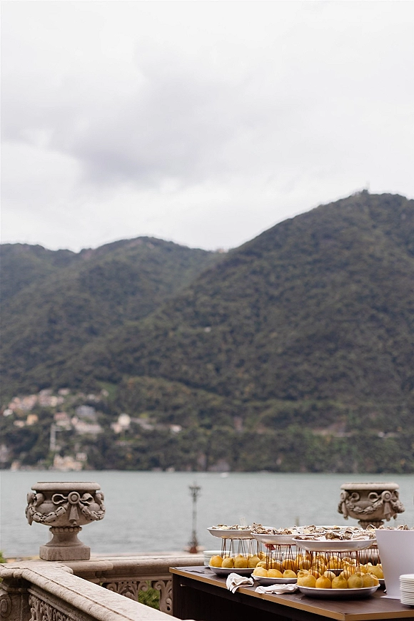 Wedding cocktail hour buffet table with oysters and lemon garnish on elevated stands, set on a stone terrace overlooking a mountain lake