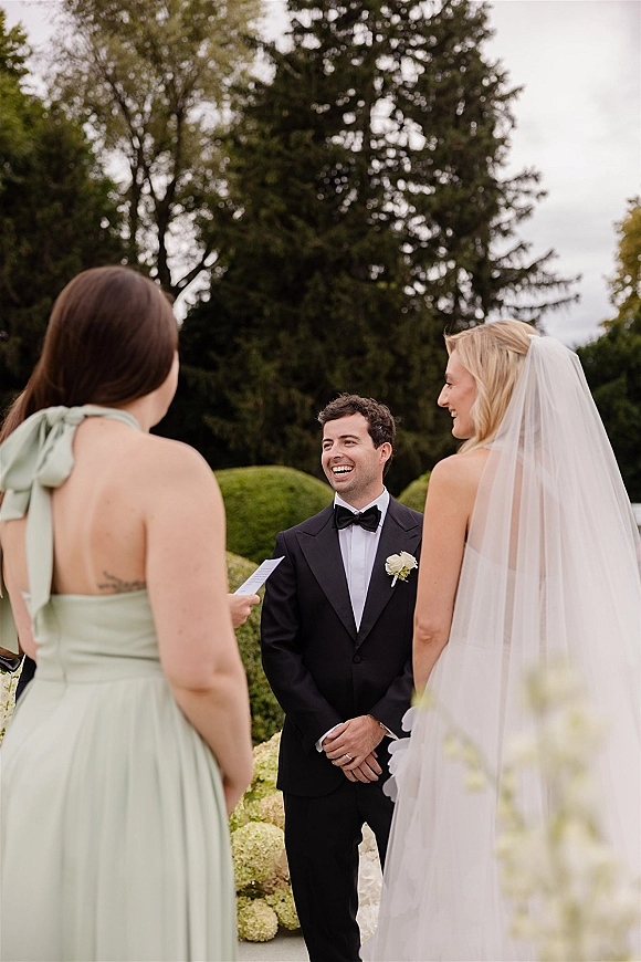 Wedding vows as the groom reacts while reading printed vows beside the bride with a long veil, in a cloudy garden ceremony setting