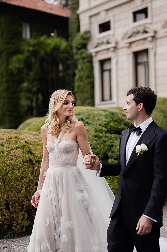 Couple portrait of bride and groom walking hand in hand, bride in veil and strapless gown beside groom in black tuxedo on garden path