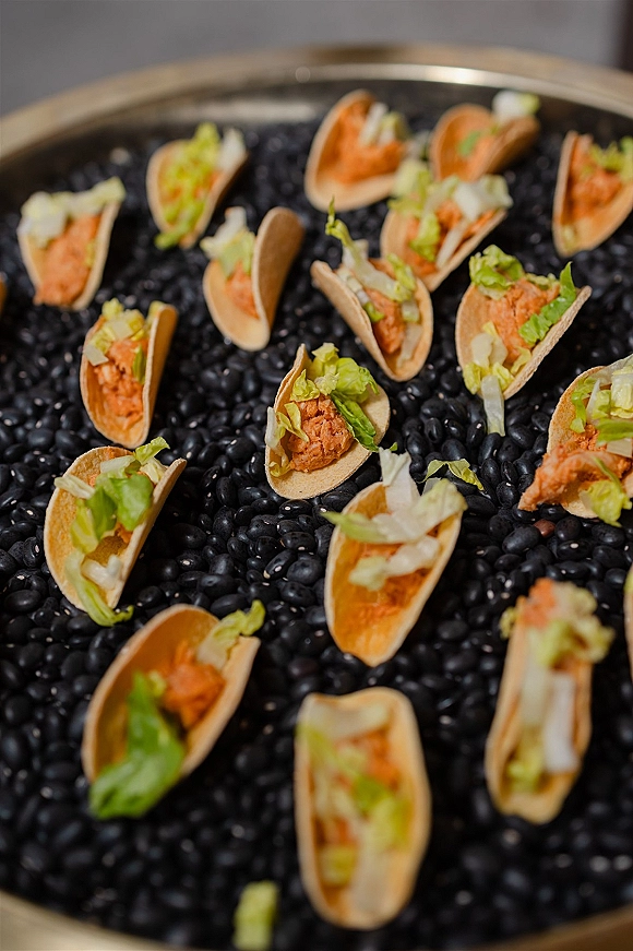 Wedding appetizers featuring mini taco bites topped with black beans, lettuce, and diced onion arranged on a serving tray on a tabletop display