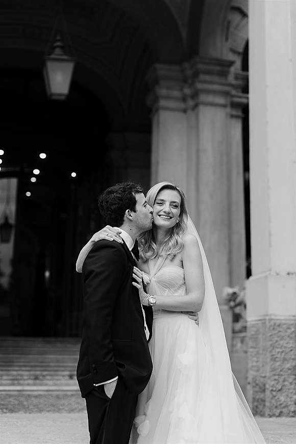 Wedding couple portrait of groom kissing bride’s cheek as she smiles in a veil and strapless dress under a stone archway entrance