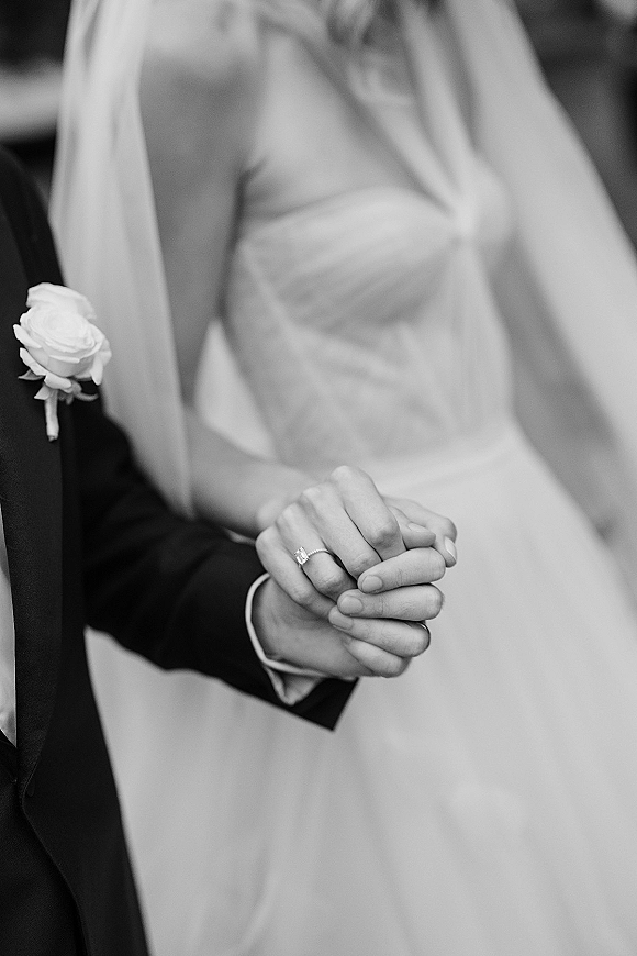 Wedding hands close-up of bride and groom holding hands, solitaire engagement ring visible, with blurred ceremony setting behind them