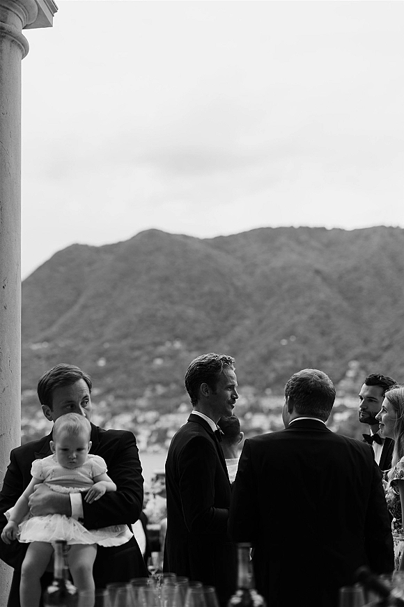 Wedding cocktail hour with wedding guests mingling in tuxedos and bow ties, a father holding a baby, on a lakeside terrace with mountains
