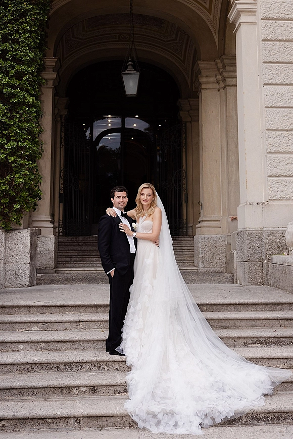 Couple portrait of bride and groom on steps, her veil and long train beside his tuxedo, framed by an ivy wall and arched doorway.
