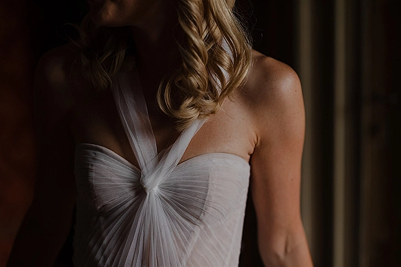 Wedding dress close-up of a strapless wedding dress with ruched bodice, tulle overlay, and knot detail in moody window light indoors