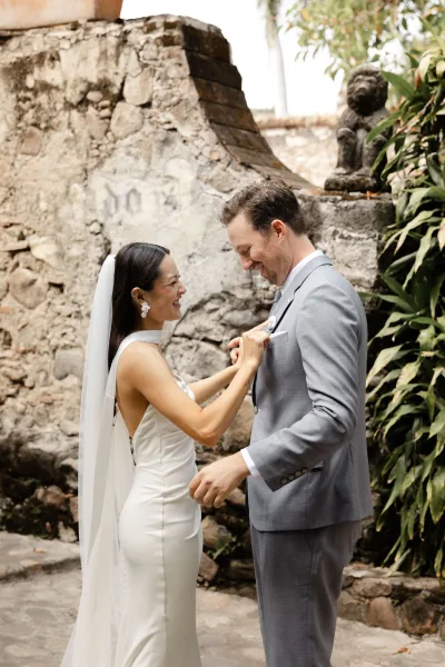 First look moment as bride fixes boutonniere on groom’s suit, both laughing beside a stone wall and greenery on an outdoor walkway