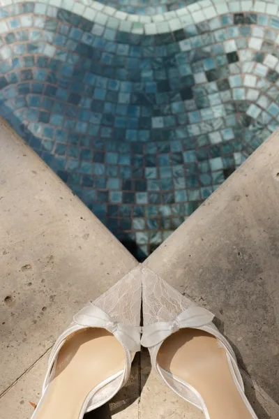 Bridal shoes, white satin heels with bows and lace detail arranged on a stone surface beside blue mosaic tile for something blue accents