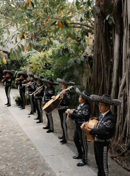 Mariachi wedding band plays trumpets and violins in embroidered charro suits with sombreros on a stone walkway amid tropical garden greenery