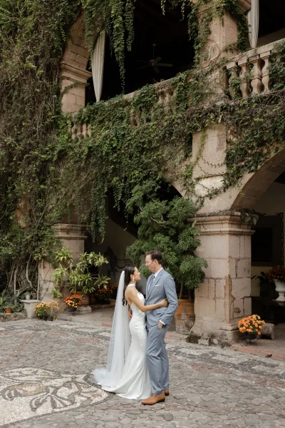 Couple portrait of bride and groom embrace, her long veil and satin dress flowing as they stand under an ivy stone archway courtyard