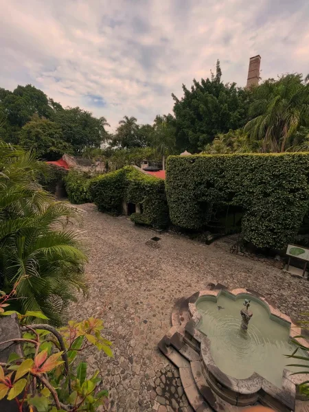 Outdoor wedding venue with a stone fountain in a cobblestone courtyard, trimmed hedges and palm trees near red roof tiles under cloudy sky