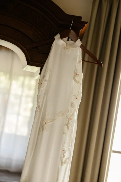 Wedding dress on hanger in window light, showing high neckline and embroidered lace appliques, framed by curtains and carved wooden headboard