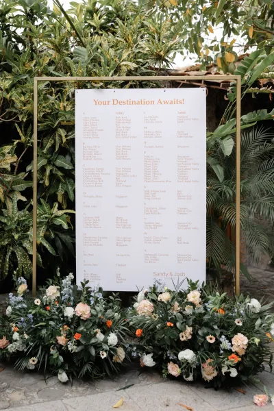 Wedding seating chart in a gold frame with roses, hydrangea, and greenery, displayed on a stone patio amid tropical plants