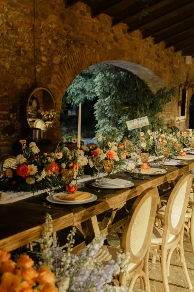 Reception tablescape on a wood farm table with floral garland, orange roses, tall taper candles, and amber goblets in a stone arch venue