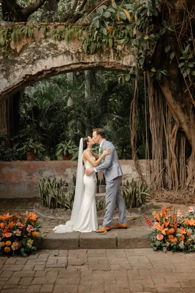 Wedding kiss portrait of bride and groom kissing in a sleek gown and gray suit, long veil flowing on stone steps in a tropical courtyard under hanging roots