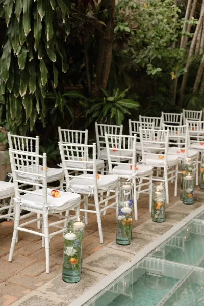 Ceremony aisle decor with white chiavari chairs, ribbon ties, and rose petals beside glass cylinder vases with floating candles by poolside greenery