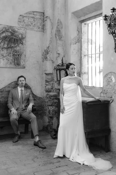 Couple portrait in black and white wedding portrait style, bride in strapless gown with train beside groom on a bench by a barred window