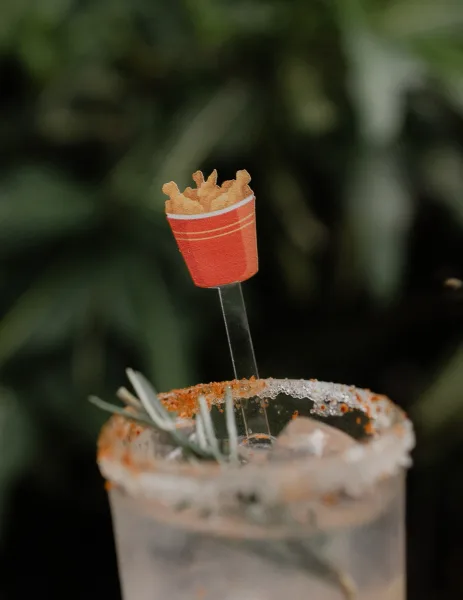 Wedding cocktail with a chili salt rim, ice, and rosemary sprig, finished with a novelty stirrer and french fry garnish against blurred greenery