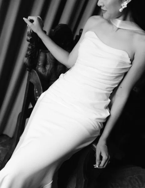 Bridal portrait in black and white of a bride in a strapless wedding dress, pearl drop earrings and floral hair clip, seated by draped curtains