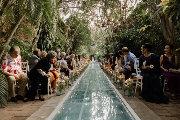 Ceremony aisle design with a glass aisle runner lined by candles in cylinder vases and low florals, set on a stone walkway in a tropical garden.