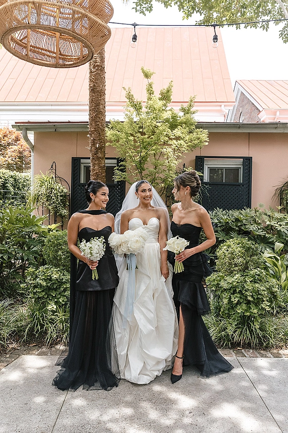 Bride with bridesmaids in black dresses holding white rose bouquets, bride in strapless gown and veil under string lights in a garden patio setting