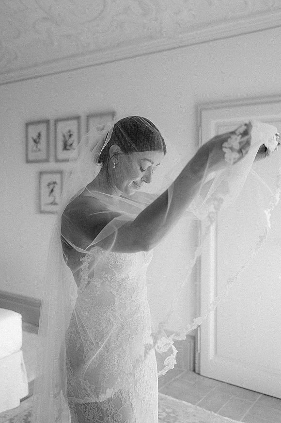 Bride getting ready, bride putting on veil over her face in a strapless lace gown with drop earrings in a quiet bedroom setting