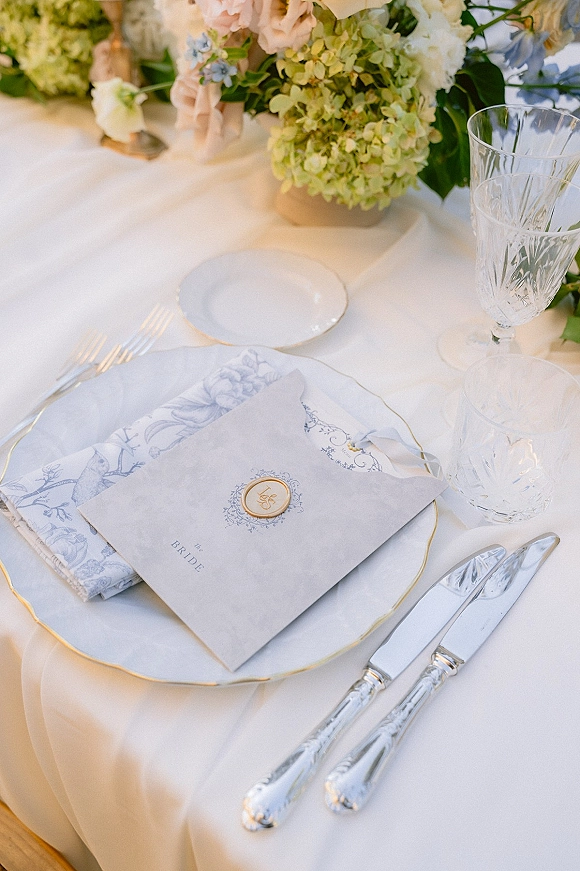 Wedding place setting with gold rim plate, printed napkin and wax seal menu card, crystal goblets, silver flatware on white tablecloth in soft light