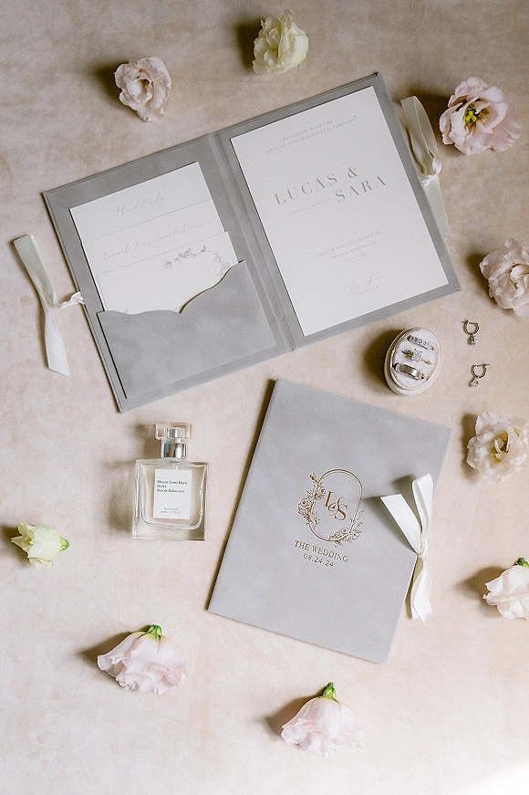 Wedding invitation suite with gray wedding invitations in a folder, silk ribbon, monogram crest, rings and blush flowers on linen backdrop