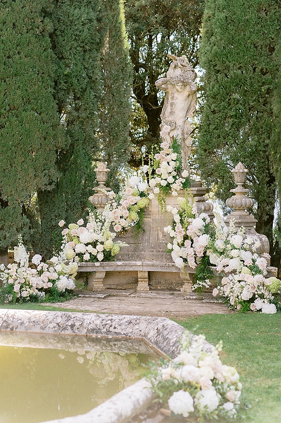 Ceremony altar with garden wedding altar florals, stone statue and bench, rose and hydrangea arrangements beside a fountain pool lawn