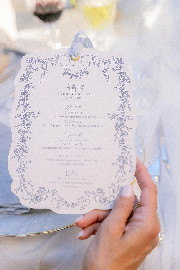 Wedding menu card with blue floral design and scalloped edge, tied with ribbon on a white plate place setting, hand with rings nearby