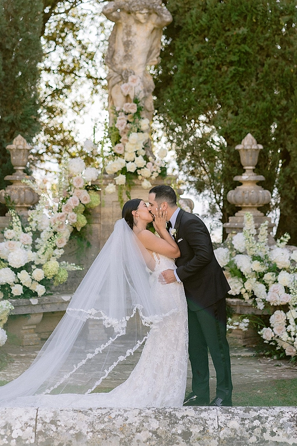 Wedding kiss portrait of bride and groom kissing under a white and blush rose floral arch on a stone terrace by a fountain