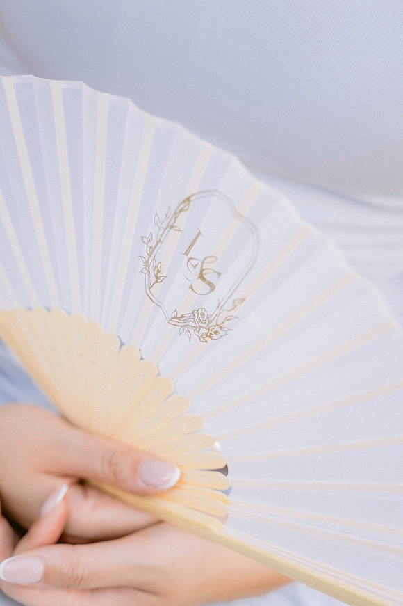 Wedding hand fan with personalized wedding fan monogram and floral crest on a paper fan with wooden handle, held over soft fabric backdrop