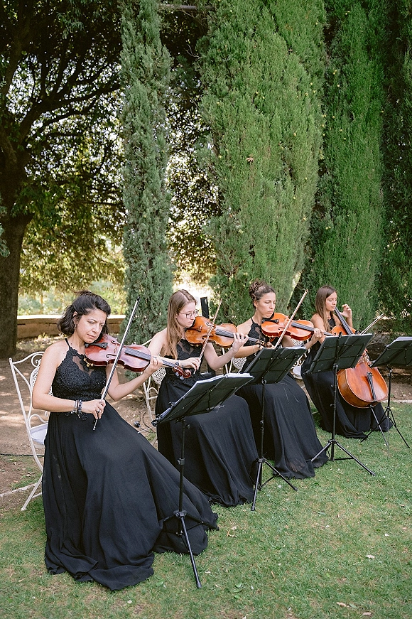 Wedding musicians performing string quartet wedding music with violins and cello, reading sheet music on stands on a garden lawn under trees