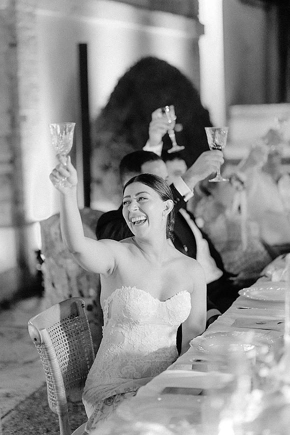 Wedding toast as the bride gives toast, laughing in a strapless lace dress with raised champagne flute at a stone-walled reception hall