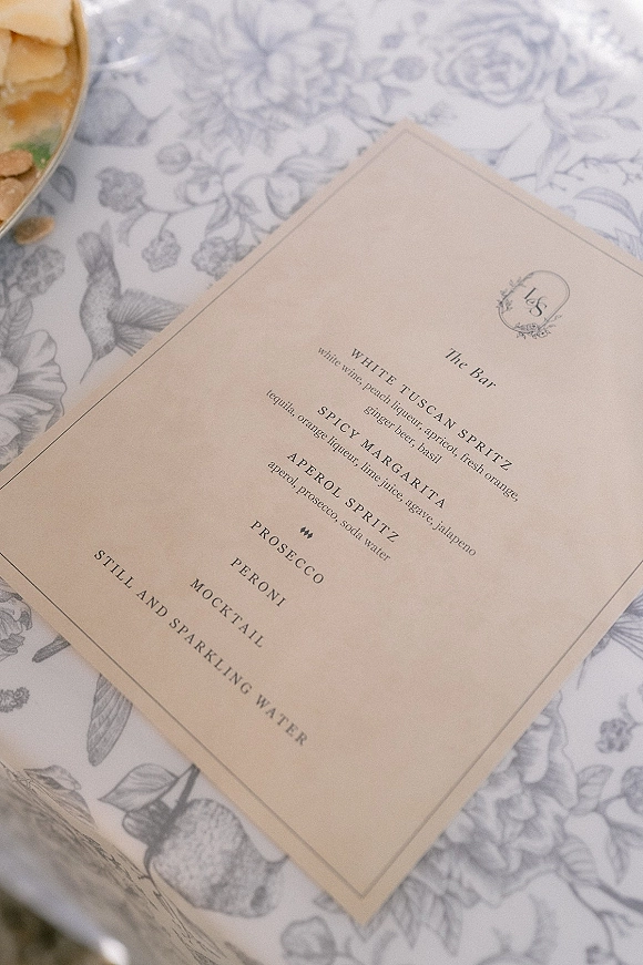Wedding bar menu card with classic serif monogram and drink list on blue-and-white toile tablecloth beside a garnish bowl