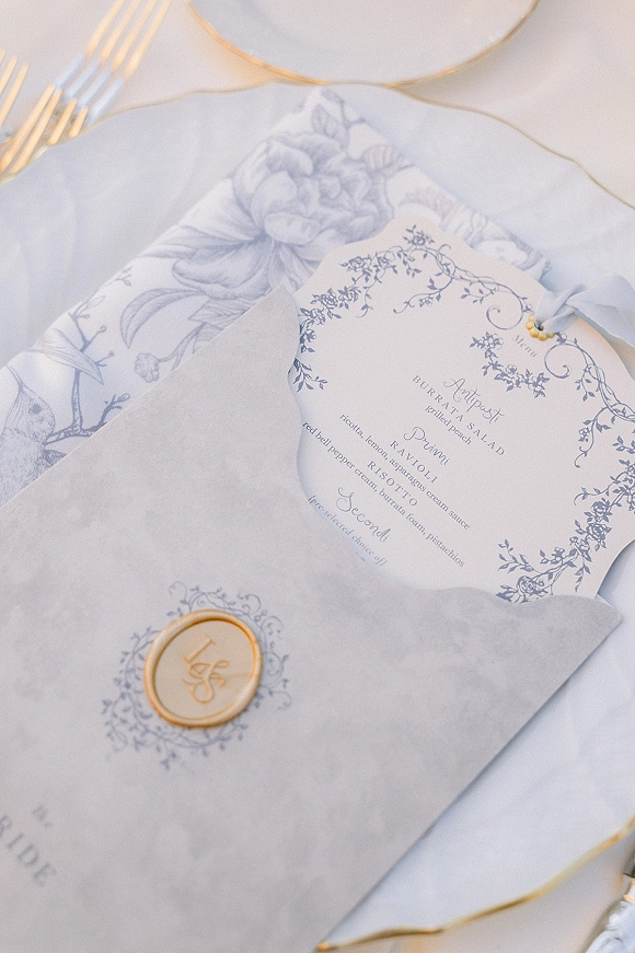 Wedding invitation suite with blue and white wedding invitations, wax seal envelope and menu card on white linen with gold-rimmed plates