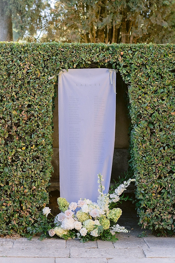 Wedding seating chart on a fabric seating chart banner with calligraphy and ribbon ties, framed by roses and hydrangea on a hedge wall