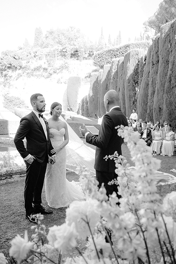 Wedding vows as bride and groom hold hands with officiant, in an outdoor wedding ceremony garden with seated guests and water feature