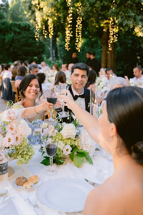 Wedding reception toast with bride and groom clinking crystal glasses at a long garden table under string lights, roses and hydrangea centerpiece