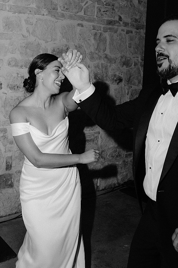 Wedding dance moment as bride in off-the-shoulder dress twirls with groom in tuxedo and bow tie on an indoor dance floor by a stone wall