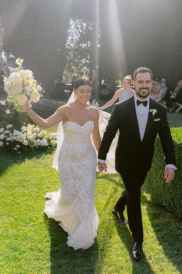 Wedding recessional as bride and groom walk the aisle holding hands, bride raising bouquet, backlit on a sunny garden lawn with guests