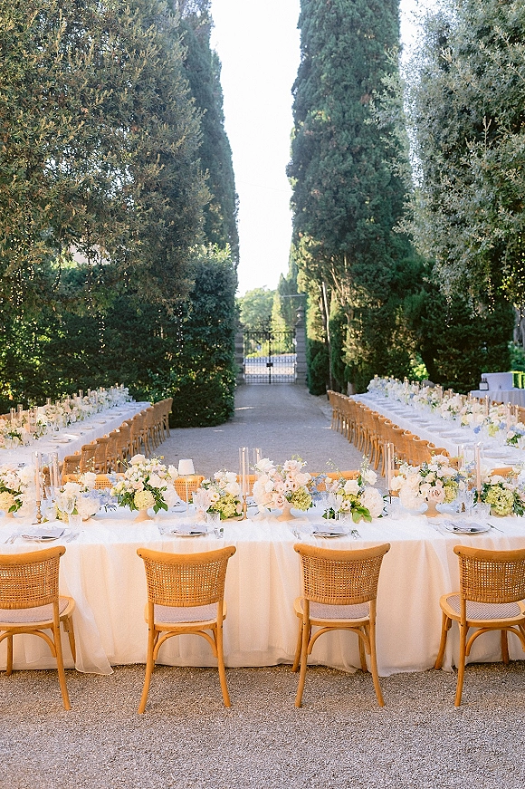 Outdoor reception tablescape with long banquet table setup, white linens, rattan chairs, pastel florals and taper candles under string lights in a cypress-lined path