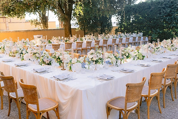 Reception tablescape for an outdoor wedding reception with long white linen banquet tables, low florals, candles, rattan chairs, and string lights overhead