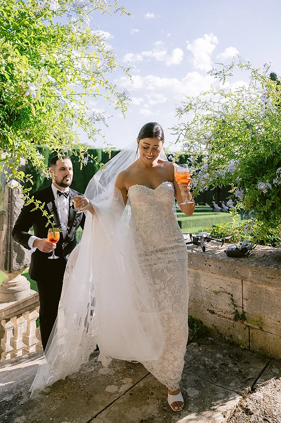 Wedding couple portrait of bride in lace dress and long veil with groom in tuxedo, holding orange cocktails on a stone terrace garden staircase