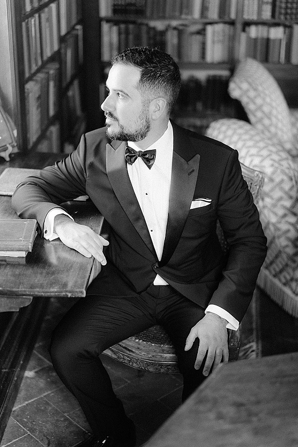 Groom portrait in a black tuxedo groom look with bow tie, seated by a wooden table in a library, lit by soft window light