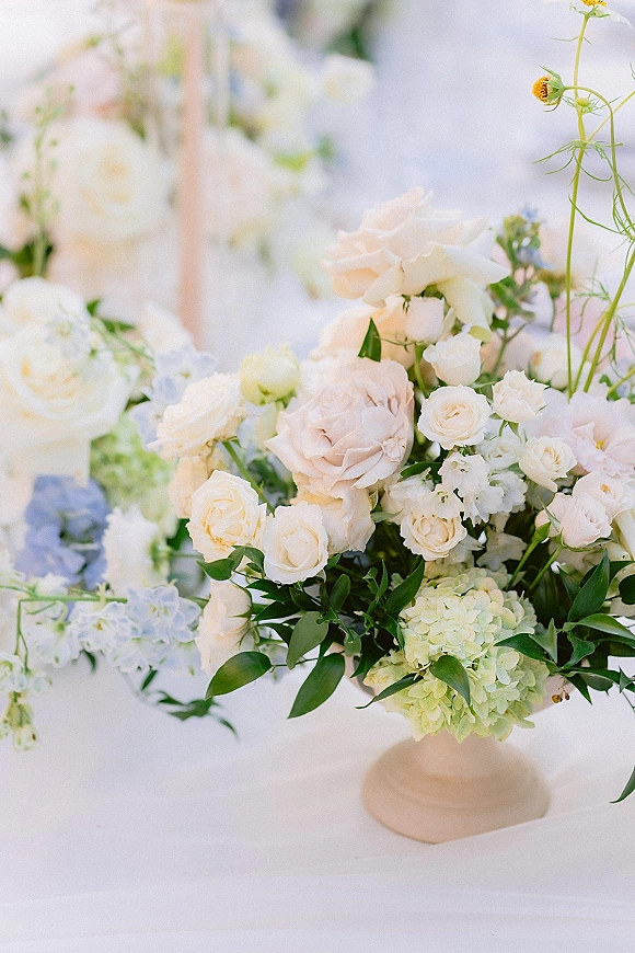 Wedding centerpiece with white rose accents featuring blush roses, hydrangea, baby’s breath, and greenery in a compote vase on linens