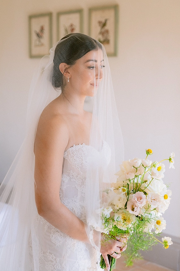 Bridal portrait of a bride with veil in side profile, holding a blush rose bouquet in soft natural light beside framed artwork