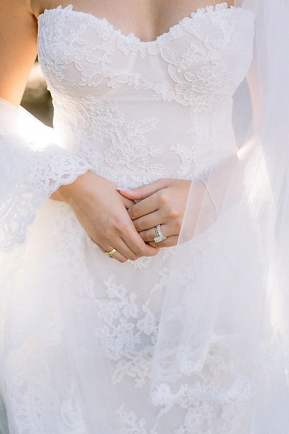 Wedding dress close-up showing a strapless lace wedding dress bodice, bride’s hands with diamond ring and veil in soft natural light