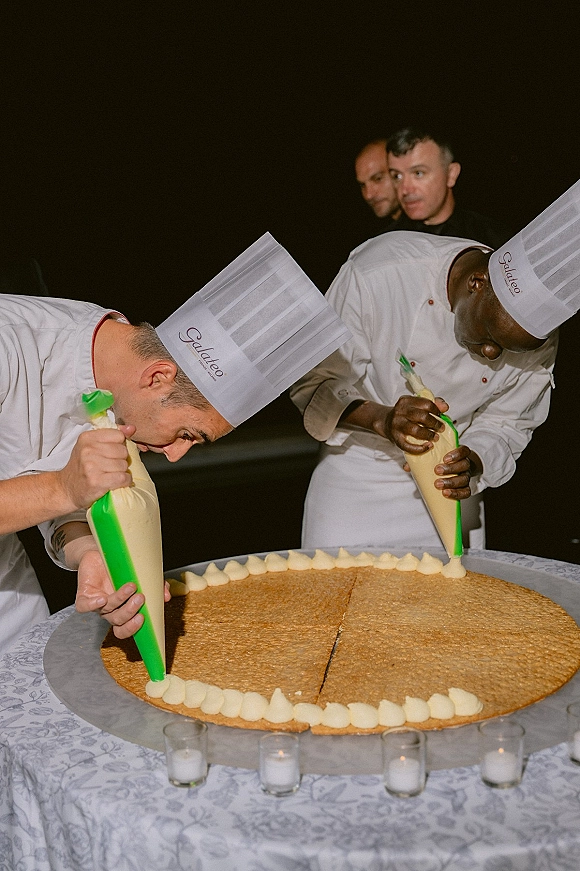 Wedding cake decorating by pastry chefs at wedding reception, piping cream frosting onto a giant round cake on a candlelit table