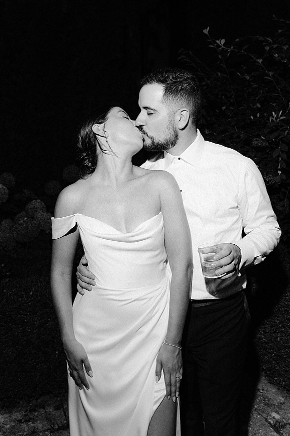 Wedding kiss portrait in black and white of bride and groom kissing at night, her off-shoulder slit gown and his drink against garden foliage