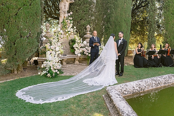 Wedding ceremony moment as bride and groom hold hands under a floral arch, lace veil train flowing, string quartet by a garden fountain